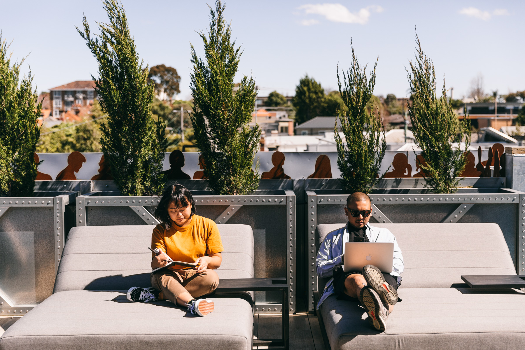 Relaxed rooftop workspace at Deskplex Office Space Hawthorn Richmond Abbotsford with two people, one holding a notebook and the other using a laptop, surrounded by tall green plants