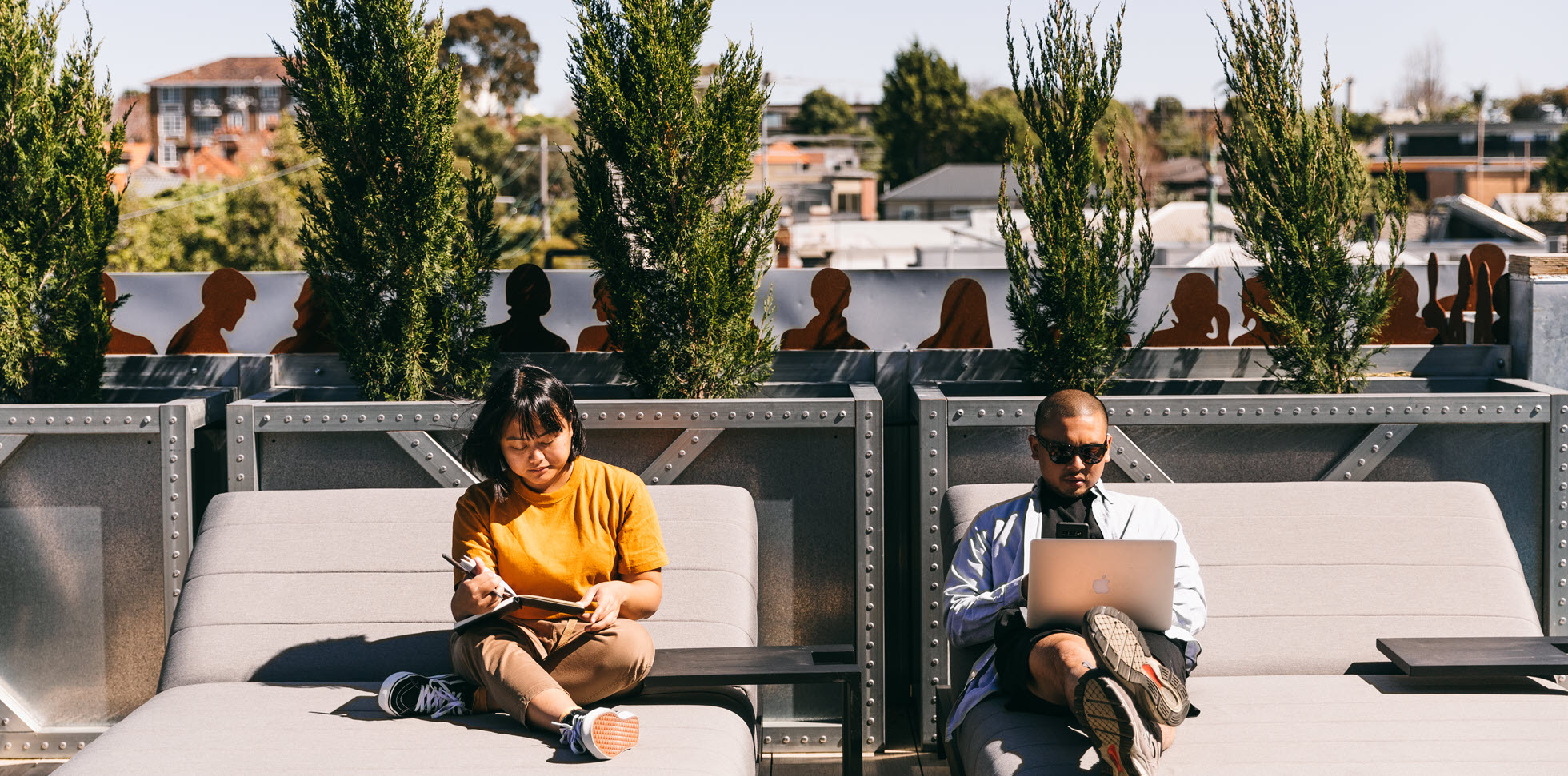 Relaxed rooftop workspace at Deskplex Office Space Hawthorn Richmond Abbotsford with two people, one holding a notebook and the other using a laptop, surrounded by tall green plants