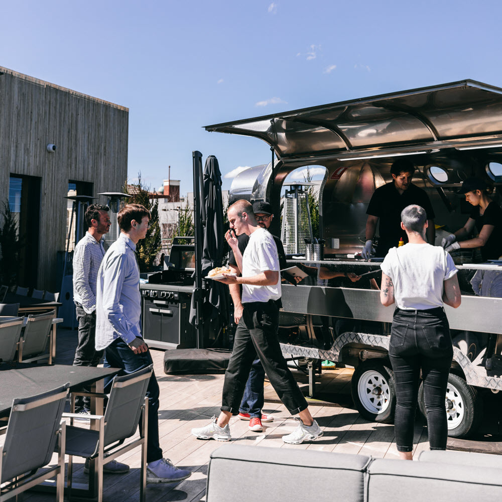 Outdoor food truck at Deskplex Office Space Hawthorn Richmond Abbotsford with a shiny metallic exterior, serving food to a group of people enjoying a sunny day in a casual, social atmosphere. Includes seating arrangements with tables and chairs