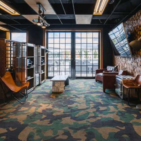 Modern, industrial-style shared office space with dark walls and a camouflage carpet at DeskPlex Office Space Hawthorn Richmond Abbotsford. Features wooden walls and ceiling, metal shelving, lighting fixtures, leather armchairs, large windows with black frames, a coffee table, and a fireplace, creating a comfortable and inviting workspace.