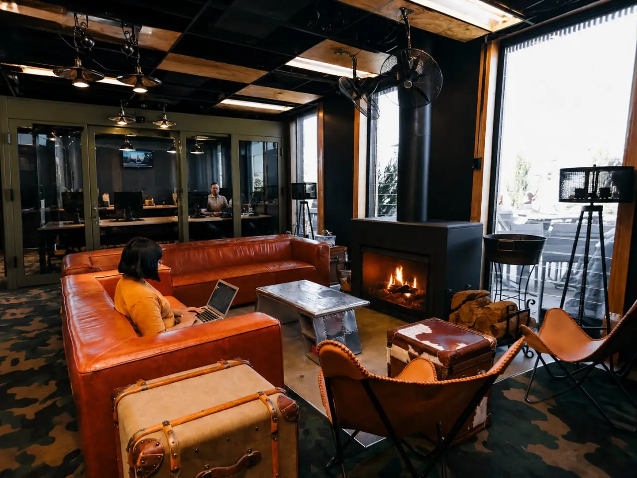 Modern coworking lounge with leather sofas, fireplace, and open office workspaces, featuring a professional remote work environment with industrial interior design.