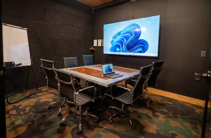 Contemporary small conference room with square meeting table, rolling office chairs, laptop setup, and large wall-mounted display in a modern coworking office.