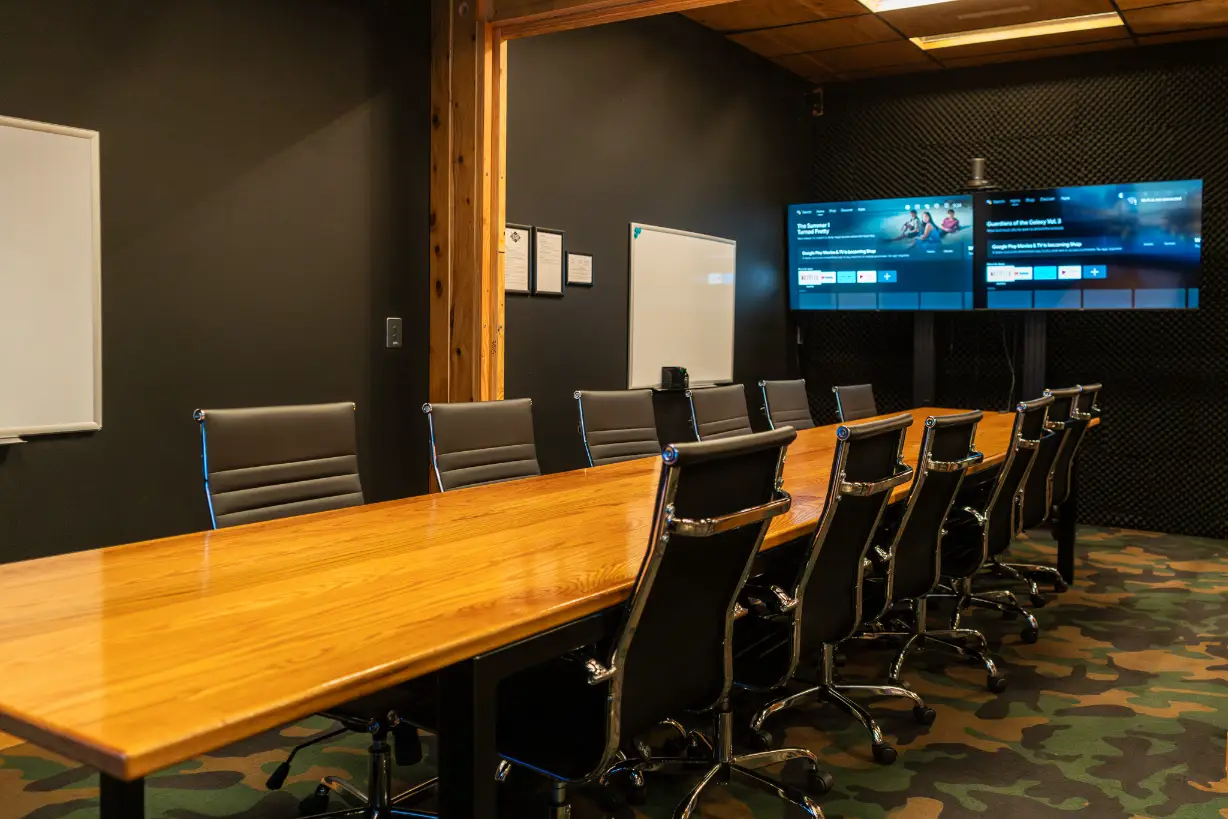 Modern executive conference room with long wooden meeting table, black leather office chairs, dual flat-screen displays, and acoustic wall panels in a professional workspace.