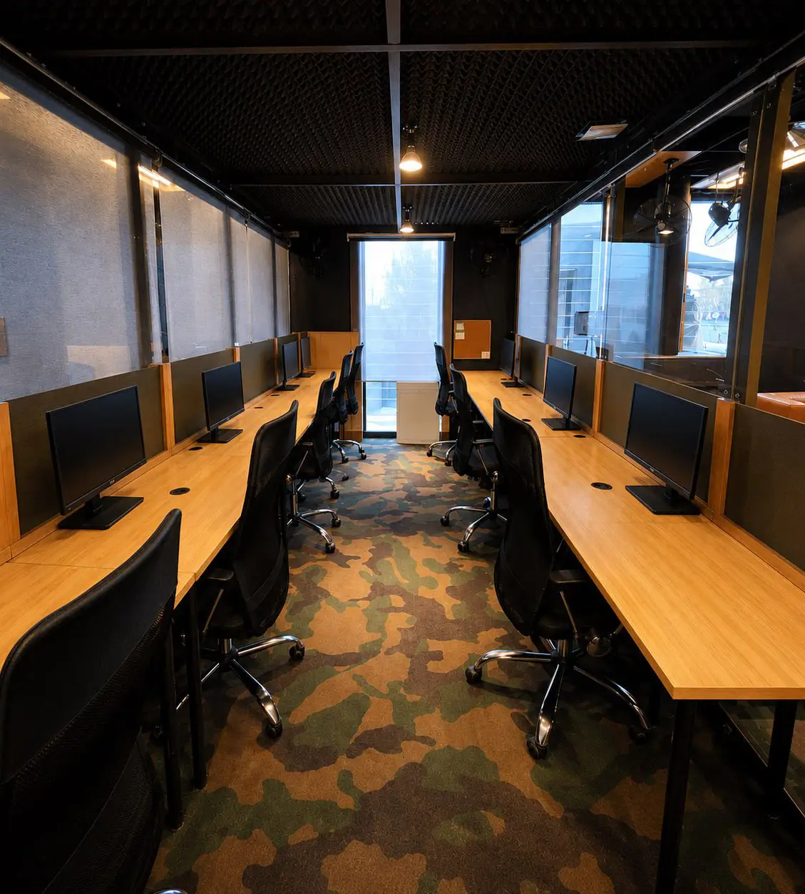 Long shared office workspace with dual-sided wooden desks, desktop monitors, ergonomic chairs, and camouflage-pattern flooring in a modern coworking office.
