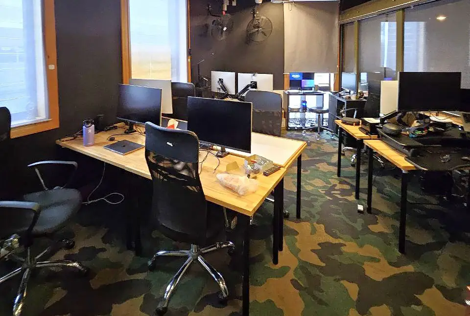 Modern shared coworking office with multiple computer workstations, ergonomic chairs, wooden desks, and camouflage-pattern flooring in a collaborative tech workspace.